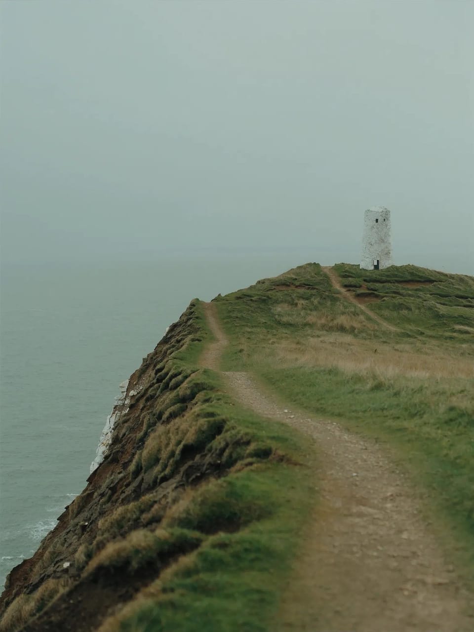 A narrow grass path along a low cliff leading to a small white tower at the headland.