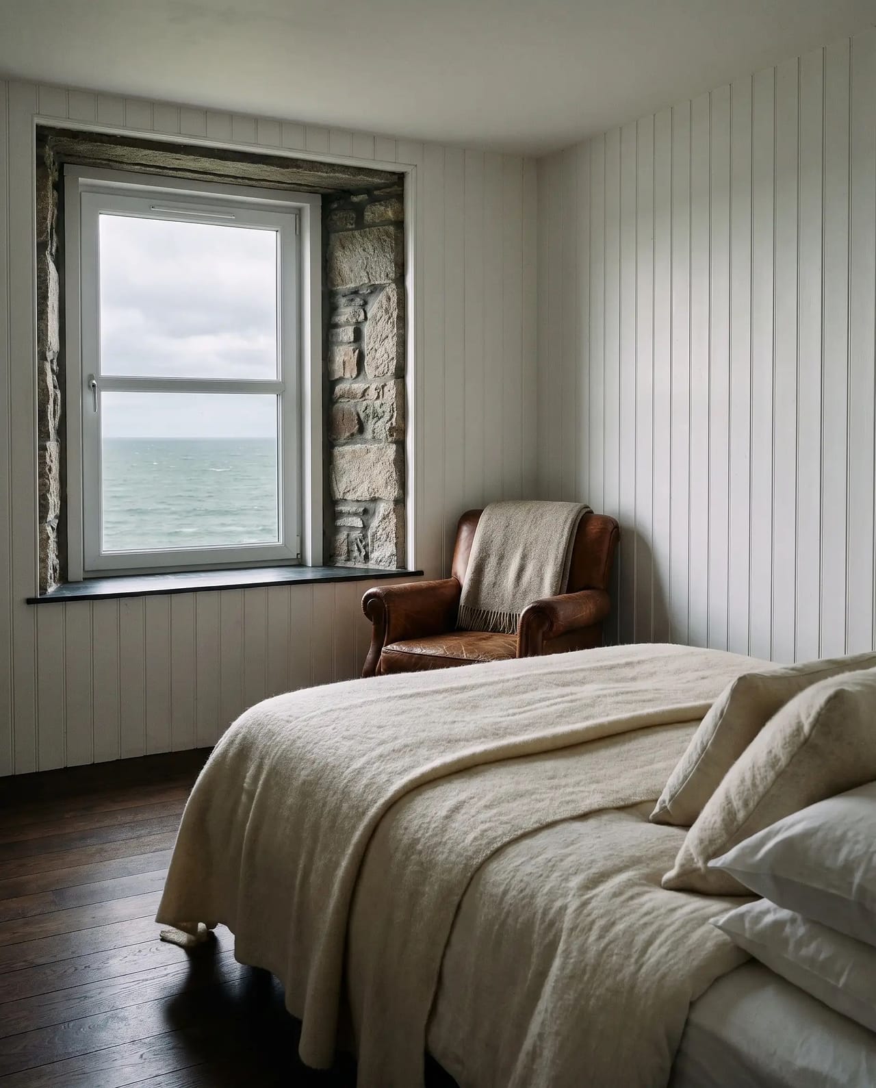 A corner bedroom with oak floorboards, a bed dressed in undyed wool and a deep-set window onto grey sea.